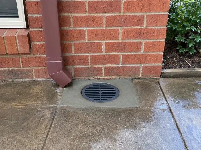Clean outdoor drain grate beside brick wall and downpipe on wet paving in Prairiewood
