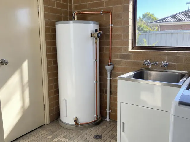 Hot water cylinder with copper pipework beside a laundry sink and brick wall in Wetherill Park